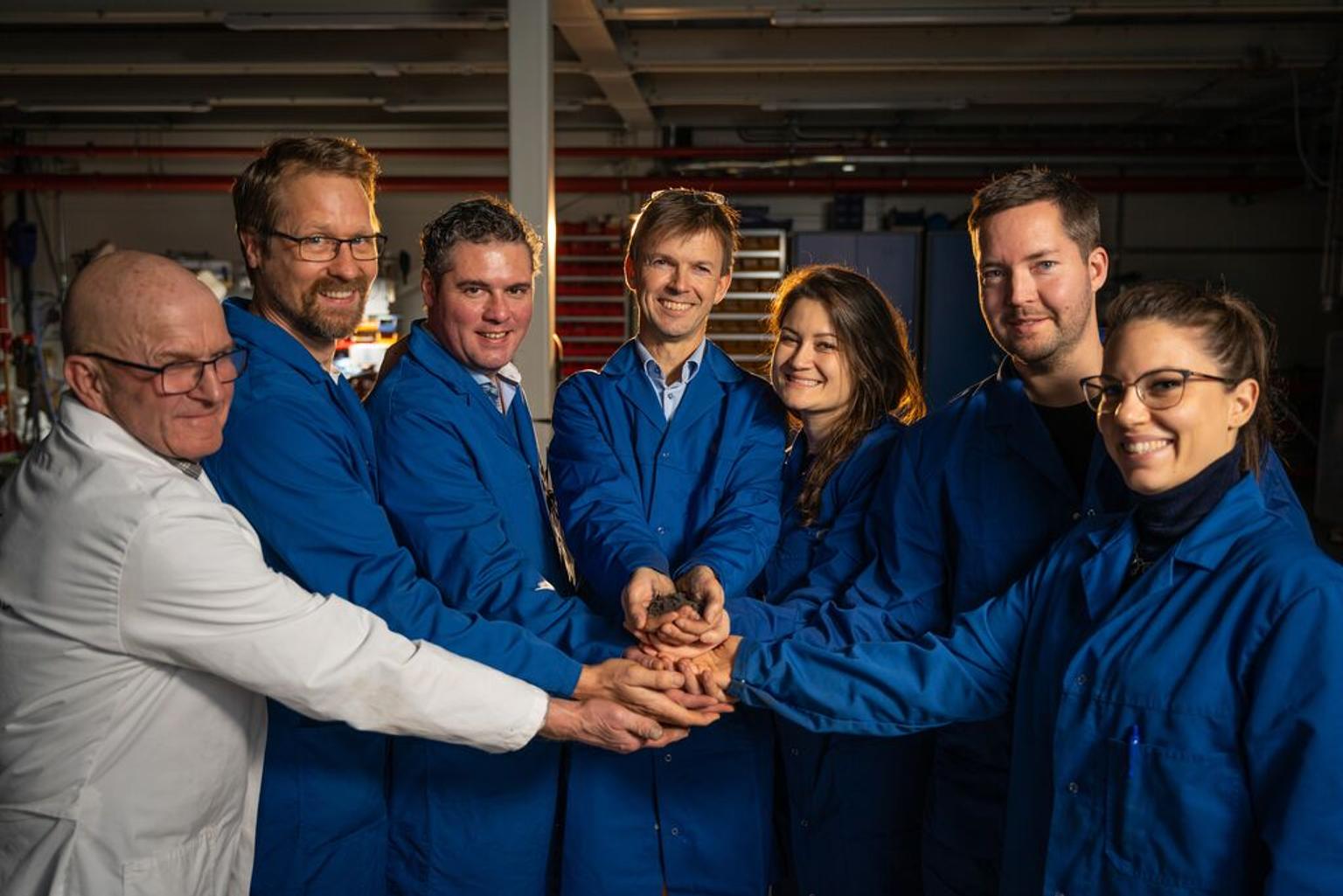 Six people in blue lab coats and one person in a white lab coat holding hands